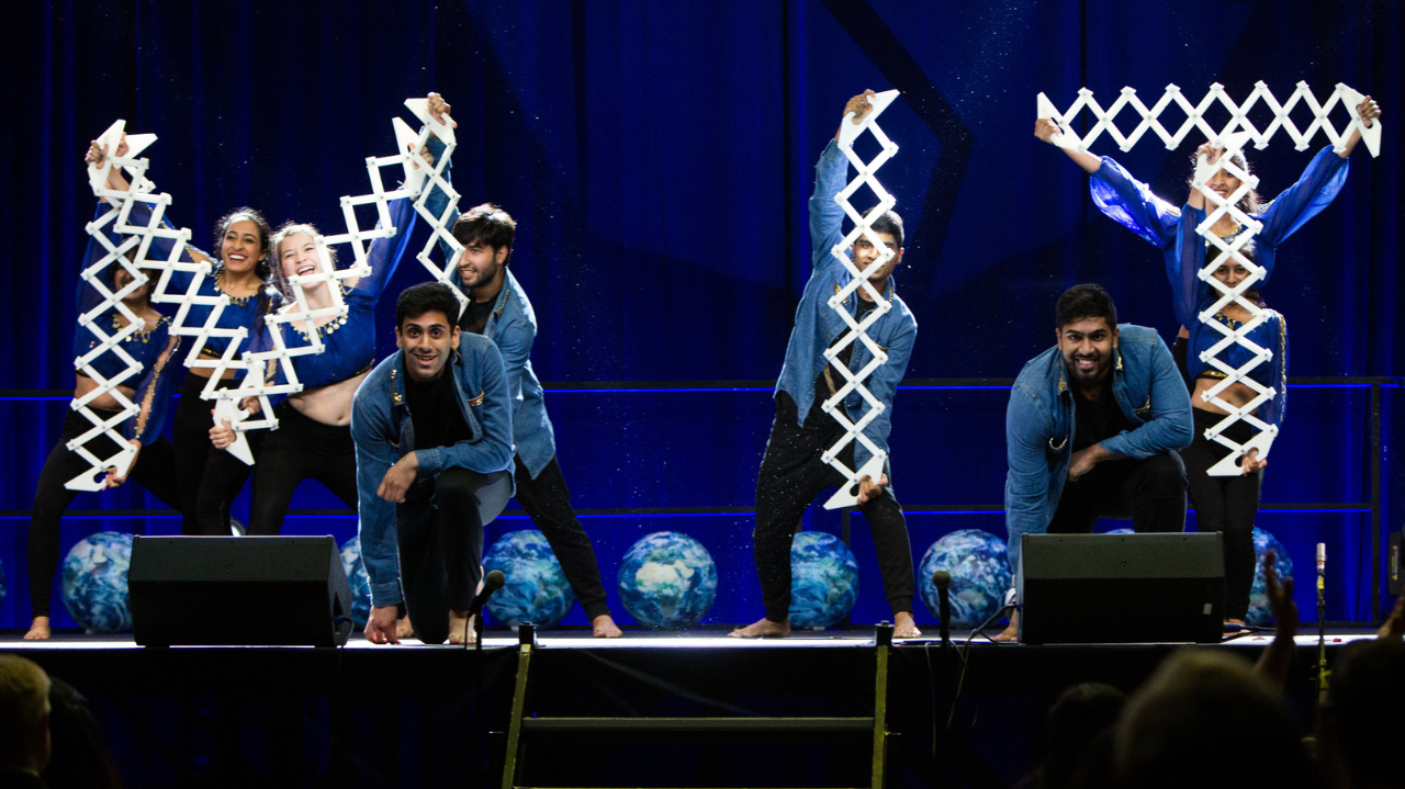 Photo of dancers on stage posing at the end of a performance, each holding large, unfoldable letters that spell out M I T